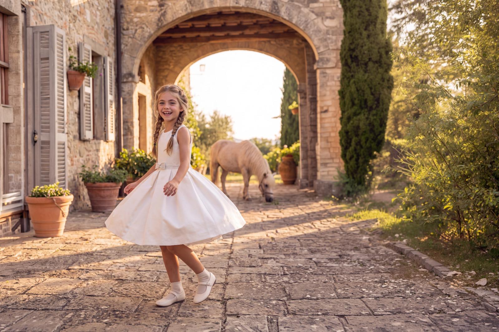 Lachend meisje in communiejurk op de kasseien voor een Toscaanse poort, met een pony onder de boog